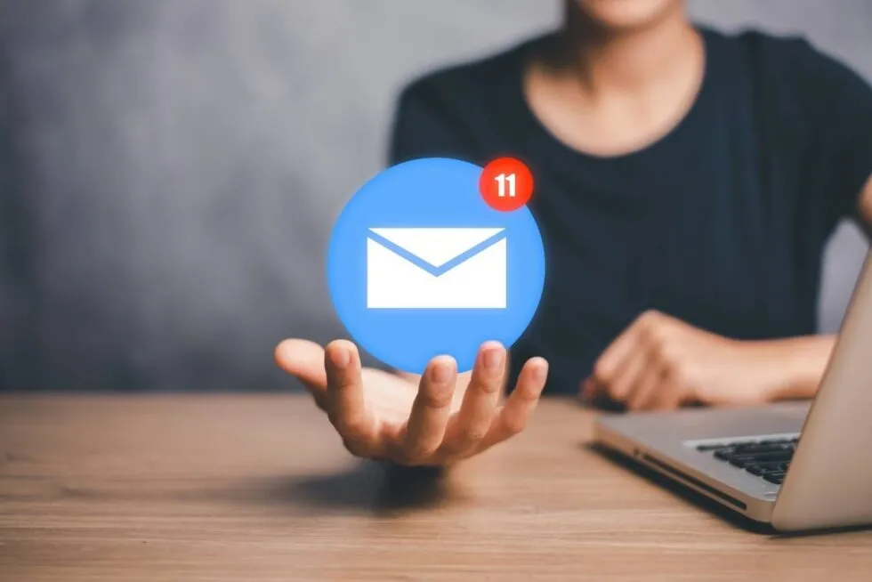 Hand holding a floating email icon with 11 unread messages, beside a laptop