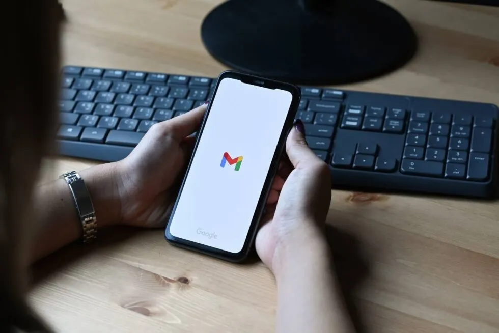 Hands holding a phone showing the Gmail logo, above a keyboard